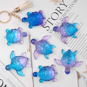 May include: A collection of translucent turtle-shaped keychains in shades of blue and purple. The keychains are arranged on a white surface with a gold-colored keychain and a tassel. The word "STORY" is visible in the background.
