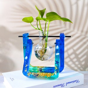 Puede incluir: Un jarrón de resina azul y verde con un bulbo de vidrio transparente para sostener una planta. El jarrón tiene una barra de metal en la parte superior y está decorado con escamas doradas y conchas.
