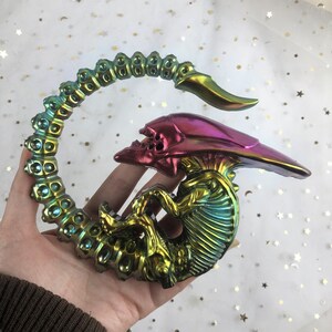 May include: An iridescent green, purple and gold alien creature figurine with a curled tail. The figurine is made of resin and has a detailed design.