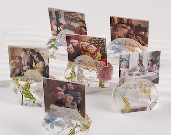 May include: Clear resin photo holders with embedded dried flowers and greenery. Each holder displays a family photo. The holders have various shapes, including round and hexagonal, and are decorated with silver glitter.