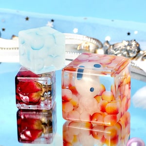 May include: Three small, clear resin cubes with different designs inside. The first cube has a white and blue swirl design. The second cube has a red rose and gold leaf design. The third cube has a white and orange bead design with a white figure inside.