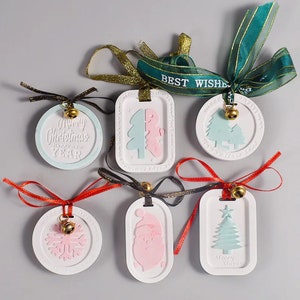 Può includere: Cinque ornamenti natalizi in ceramica bianca con diversi design. Gli ornamenti sono decorati con vernice rosa e blu e hanno una campana attaccata a ciascuno. Gli ornamenti sono appesi a nastri di vari colori, tra cui rosso, nero e verde.