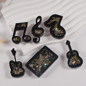 Pode incluir: Cinco pingentes de resina preta com detalhes em glitter dourado. Os pingentes têm a forma de uma clave de sol, uma nota musical, uma clave de fá, uma guitarra e uma clave de sol.