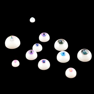 Puede incluir: Surtido de ojos de mu&ntilde;eca de pl&aacute;stico blanco con iris de varios colores, incluyendo azul, verde, morado e iridiscente.