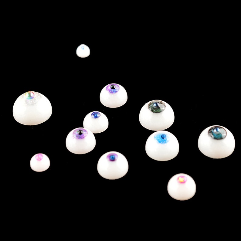 Puede incluir: Surtido de ojos de mu&ntilde;eca de pl&aacute;stico blanco con iris de varios colores, incluyendo azul, verde, morado e iridiscente.