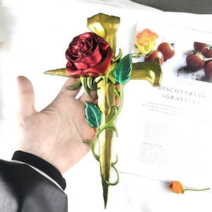 Puede incluir: Una cruz de metal de color dorado con una rosa roja y hojas verdes. La cruz tiene forma de daga con una punta puntiaguda. La rosa está en plena floración y tiene un tallo delicado. Las hojas son verdes y tienen un acabado brillante.