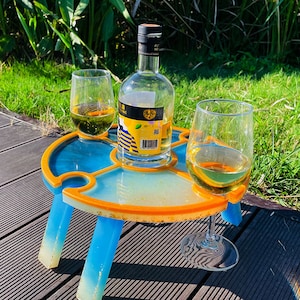 May include: A blue and yellow portable table with three legs, designed to hold two glasses and a bottle. The table is on a wooden deck with green grass in the background.