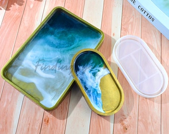 Puede incluir: Dos bandejas de resina con un diseño abstracto azul, verde y blanco. La bandeja más grande es rectangular y la más pequeña es ovalada. Ambas bandejas están sobre una superficie de madera.