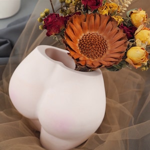 May include: White ceramic vase shaped like a human body, with a dried flower arrangement in shades of orange, yellow, and red.