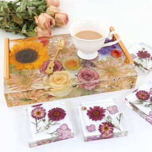 May include: A clear tray with a gold handle filled with pressed flowers, including a sunflower, roses, and daisies. Four matching coasters with pressed flowers in shades of purple and pink.