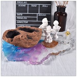 May include: A decorative tray with a blue, purple, and white resin design. A brown, textured planter sits on the tray, along with small white statues and a clear perfume bottle. A clapperboard is in the background.
