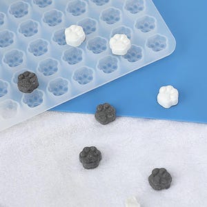 Könnte beinhalten: Eine weiße Silikonform mit Pfotenabdruckformen. Die Form ist mit kleinen, grauen und weißen Pfotenabdruck-förmigen Süßigkeiten gefüllt.