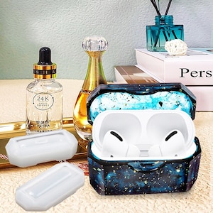 Puede incluir: Una funda protectora para AirPods de color azul y dorado con temática de galaxia, con un interior blanco y dos AirPods blancos en su interior. La funda está abierta y los AirPods son visibles. También se ven dos fundas de silicona blancas en la imagen.