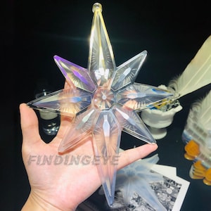 Pode incluir: Um enfeite de estrela de seis pontas transparente e iridescente com um design facetado. A estrela é segurada na mão.