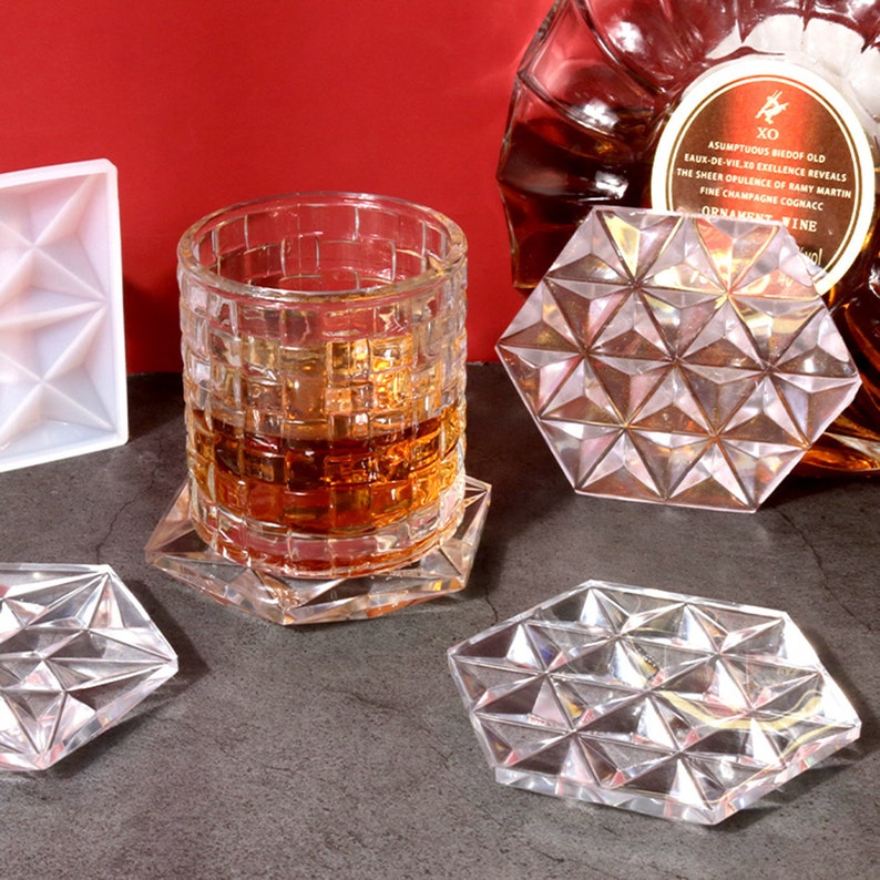 May include: Four clear glass coasters with a geometric design. The coasters are hexagonal and have a raised, diamond-like pattern. A glass of amber liquid sits on one of the coasters.