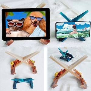 May include: Two phone stands made of clear resin with a cross design. One stand is blue and the other is brown with dried flowers. The stands are designed to hold a phone or tablet upright.