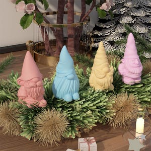 Pode incluir: Quatro gnomos coloridos com barbas brancas e chapéus pontiagudos. Os gnomos são rosa, azul, amarelo e rosa claro. Eles estão sentados em uma cama de galhos de pinheiro verdes.