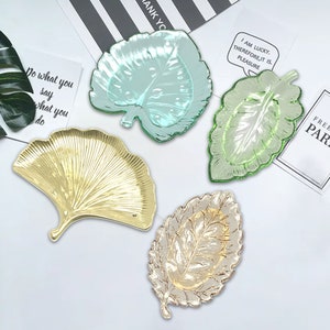 May include: Four decorative glass leaf-shaped serving trays. One tray is gold, one is light green, one is dark green, and one is a light blue-green. The trays are on a white surface.