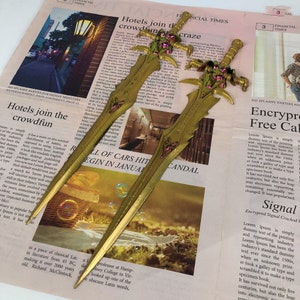 May include: Two gold-colored paper knives with intricate designs. One knife has a pink and green design, the other is a solid gold color. Both knives are lying on a newspaper with the text "Hotels join the crowdfan craze" and "Encrypro Free Call".