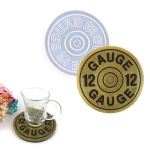 May include: A gold-colored coaster with the text "12 Gauge" printed in black. The coaster is made of a glittery material and has a round shape. A clear glass cup is sitting on top of the coaster. A white silicone mold is also shown in the image.