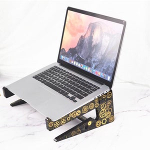 May include: A silver laptop computer rests on a black wooden stand decorated with gold-colored gears. The laptop screen displays a mountain landscape. The stand is designed to elevate the laptop for better ergonomics.