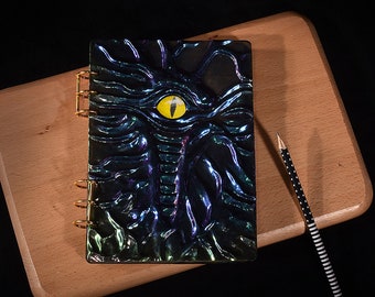 Può includere: Un quaderno nero a spirale con una copertina testurizzata che presenta un grande occhio giallo e squame iridescenti. Il quaderno è su una superficie di legno.