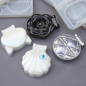 Puede incluir: Cuatro moldes de silicona transparente para espejo compacto. Los moldes tienen forma de concha, rosa, cara de gato y diseño geométrico. Los moldes están sobre una superficie gris.