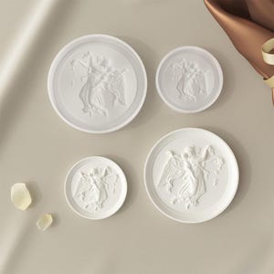 May include: Four white ceramic coasters with raised relief designs of a winged figure. The coasters are round and have a smooth, glossy finish.
