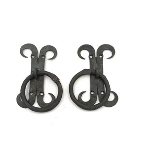 May include: Two antique-style, wrought iron door pulls. Each pull features a decorative top with curved accents, a central mounting plate, and a circular handle. The metal has a dark, aged finish, suggesting a vintage or rustic aesthetic.