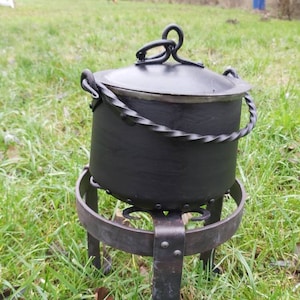 Puede incluir: Una olla de cocina de hierro fundido negro con tapa y asa de metal retorcido. La olla se asienta sobre un soporte de hierro forjado con tres patas. La olla está diseñada para cocinar al aire libre.