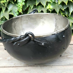 Può includere: Un calderone in metallo nero con interno argentato e costruzione rivettata. Il calderone ha due manici in metallo ritorto e piccoli piedini. L'esterno è verniciato di nero e il bordo è argentato. Questo pezzo decorativo è adatto a vari usi.