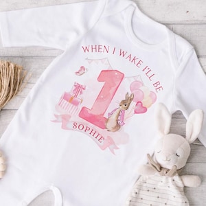May include: White baby sleepsuit with a Peter Rabbit design for a first birthday. The sleepsuit features the text "When I wake I'll be" and the name "Sophie". A stuffed bunny and wooden beads are also in the image.