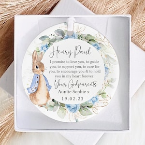 May include: A white round ornament featuring a watercolour illustration of a rabbit, flowers, and leaves. The ornament includes the text "Henry Paul" and a message about being a godparent.