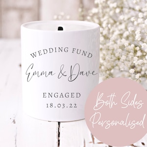May include: White ceramic jar with a slot on top for saving money. The jar is labelled "Wedding Fund Emma & Dave Engaged 18.03.22". A pink circle with the text "Both Sides Personalised" is in the foreground.