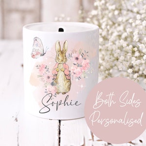 May include: White ceramic money box with a watercolour design featuring a rabbit, flowers, and a butterfly. The name "Sophie" is written on the front of the box. The text "Both Sides Personalised" is written on a pink circle.
