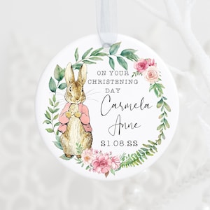 May include: A white ceramic bauble featuring a watercolour illustration of a rabbit wearing a pink jacket surrounded by green leaves and pink flowers. The text on the bauble reads "On your christening day Carmela Anne 21.08.22".