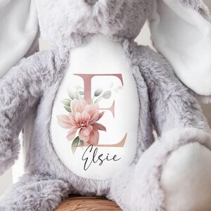 May include: A grey plush bunny with long ears sits on a wicker chair. The bunny has a white patch on its chest with a pink floral design and the letter "E" and the name "Elsie" written in cursive.