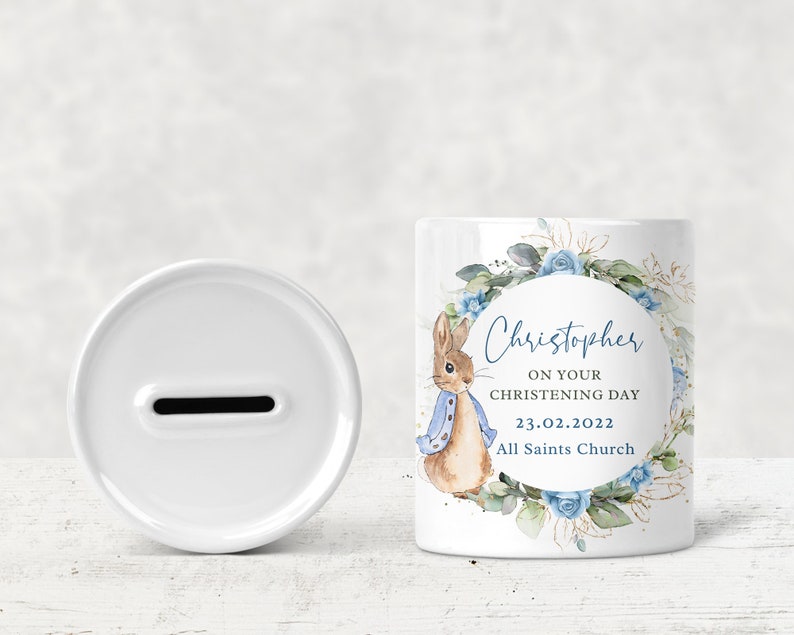May include: White ceramic money box with a blue and green floral design featuring a rabbit and the text "Christopher on your christening day 23.02.2022 All Saints Church".
