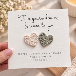 May include: A square white greeting card featuring the black text Two years down, forever to go, followed by two small crocheted hearts in cream and sage green. Below the hearts, the text reads HAPPY COTTON ANNIVERSARY, JAMES & SOPHIE, 12.08.2026.