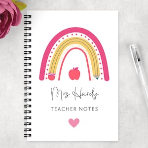 May include: Spiral-bound notebook with a white cover featuring a pink and yellow rainbow design, a red apple, and the text "Mrs Hardy TEACHER NOTES". A clear pen rests on the right. A pink flower is in the upper left corner.