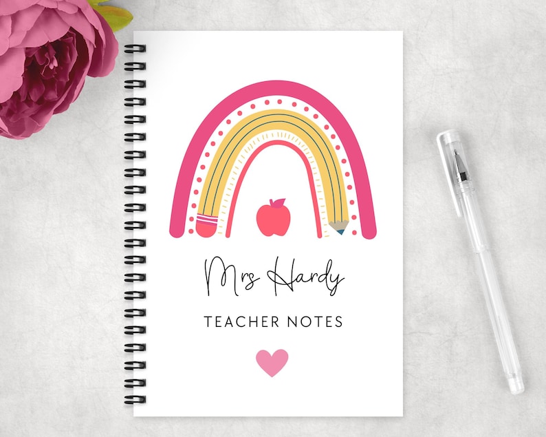 May include: Spiral-bound notebook with a white cover featuring a pink and yellow rainbow design, a red apple, and the text "Mrs Hardy TEACHER NOTES". A clear pen rests on the right. A pink flower is in the upper left corner.