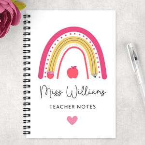 May include: A white spiral-bound notebook with a pink and yellow rainbow design, a red apple, and the text "Miss Williams TEACHER NOTES". A clear pen is on the right. A pink flower is in the upper left corner.