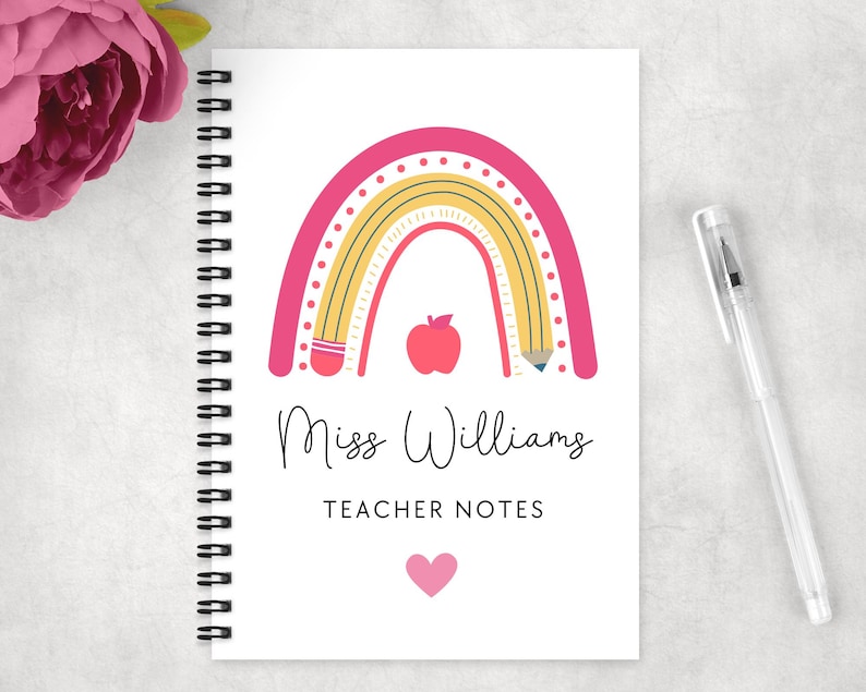 May include: A spiral-bound notebook with a white cover featuring a pink and yellow rainbow design, a red apple, and the text "Miss Williams TEACHER NOTES". A clear pen rests on the right side. A pink flower is in the upper left corner.