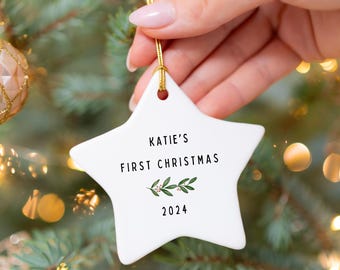 May include: White ceramic star ornament with the text "Katie's First Christmas 2024" and a small green sprig of mistletoe.