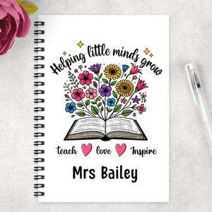 May include: A white spiral-bound notebook with the text "Helping little minds grow." The cover design features an open book with colorful flowers, the words "teach, love, inspire" and the name "Mrs Bailey."