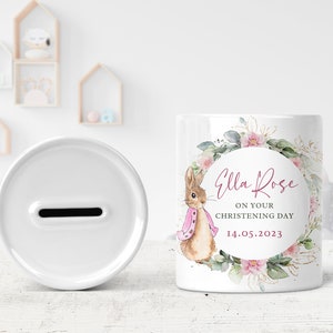 May include: White ceramic money box with a slot on the top. The front of the box features a watercolor illustration of a rabbit wearing a pink scarf, surrounded by pink flowers and greenery. The text "Ella Rose" is written in pink cursive lettering, followed by "On your christening day" and "14.05.2023".