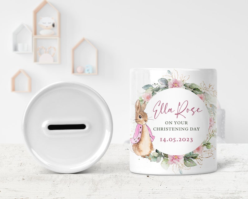 May include: White ceramic money box with a slot on the top. The front of the box features a watercolor illustration of a rabbit wearing a pink scarf, surrounded by pink flowers and greenery. The text "Ella Rose" is written in pink cursive lettering, followed by "On your christening day" and "14.05.2023".