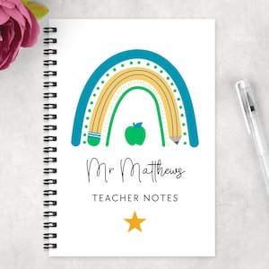 May include: A spiral-bound notebook with a white cover featuring a colorful rainbow design, a green apple, and the text "Mr Matthews TEACHER NOTES". A pen rests on the right side of the notebook. A pink flower is in the upper left corner.