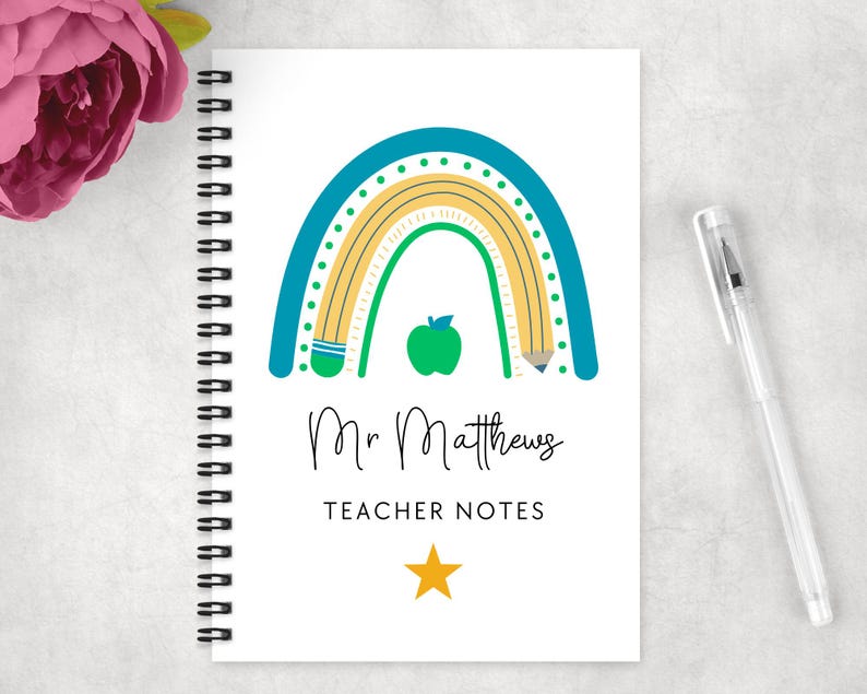 May include: A spiral-bound notebook with a white cover featuring a colorful rainbow design, a green apple, and the text "Mr Matthews TEACHER NOTES". A pen rests on the right side of the notebook. A pink flower is in the upper left corner.