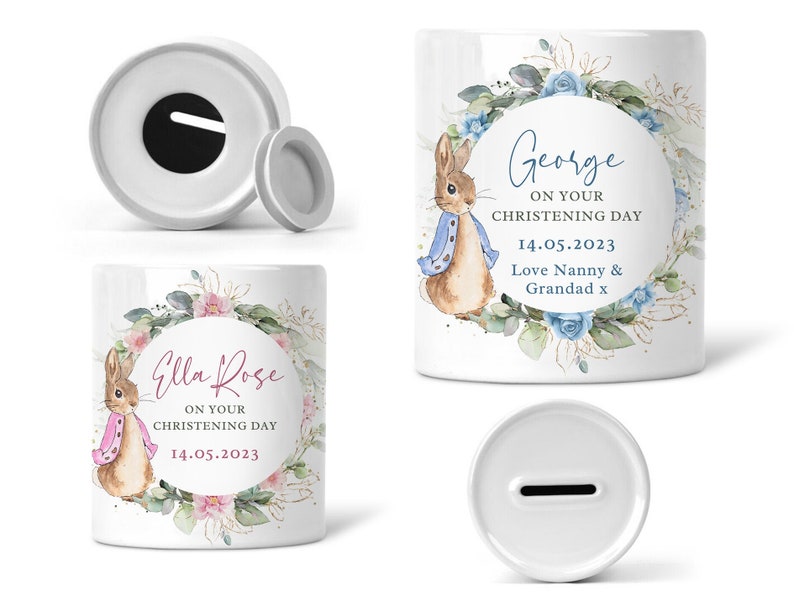 May include: White ceramic money box with a Peter Rabbit design. The box has a floral wreath with blue and pink flowers. The text on the box reads "Ella Rose on your christening day 14.05.2023".